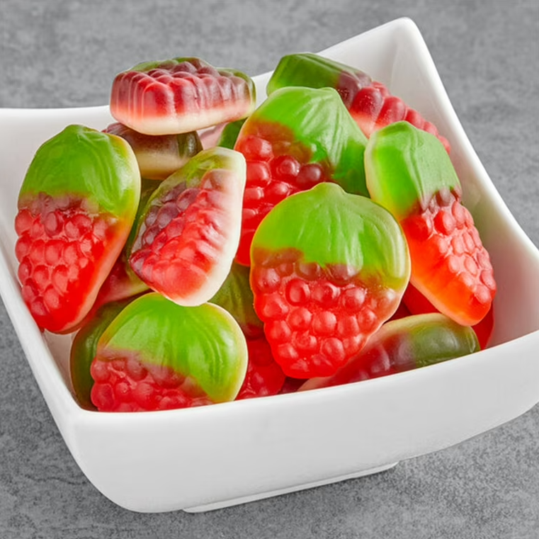 Gummy strawberry candies in red and green layered gummy shapes, arranged in a white square bowl on a gray textured background.
