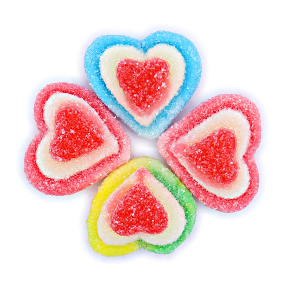 Four heart-shaped sour gummy candies arranged in a flower-like pattern on a plain white background. Each candy has a layered design with a red center, surrounded by white, and then a different outer color: blue, pink, and yellow.