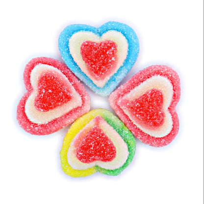 Four heart-shaped sour gummy candies arranged in a flower-like pattern on a plain white background. Each candy has a layered design with a red center, surrounded by white, and then a different outer color: blue, pink, and yellow.