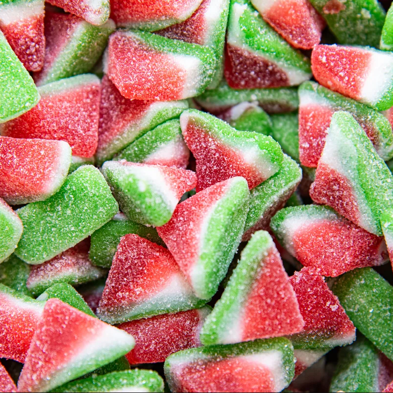 A close-up pile of sugar-dusted watermelon-shaped gummy candies, showing red flesh, green rinds, and white pith details.