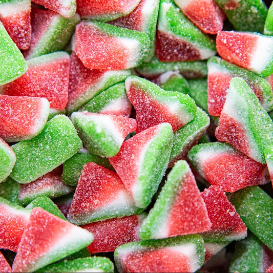 A close-up pile of sugar-dusted watermelon-shaped gummy candies, showing red flesh, green rinds, and white pith details.